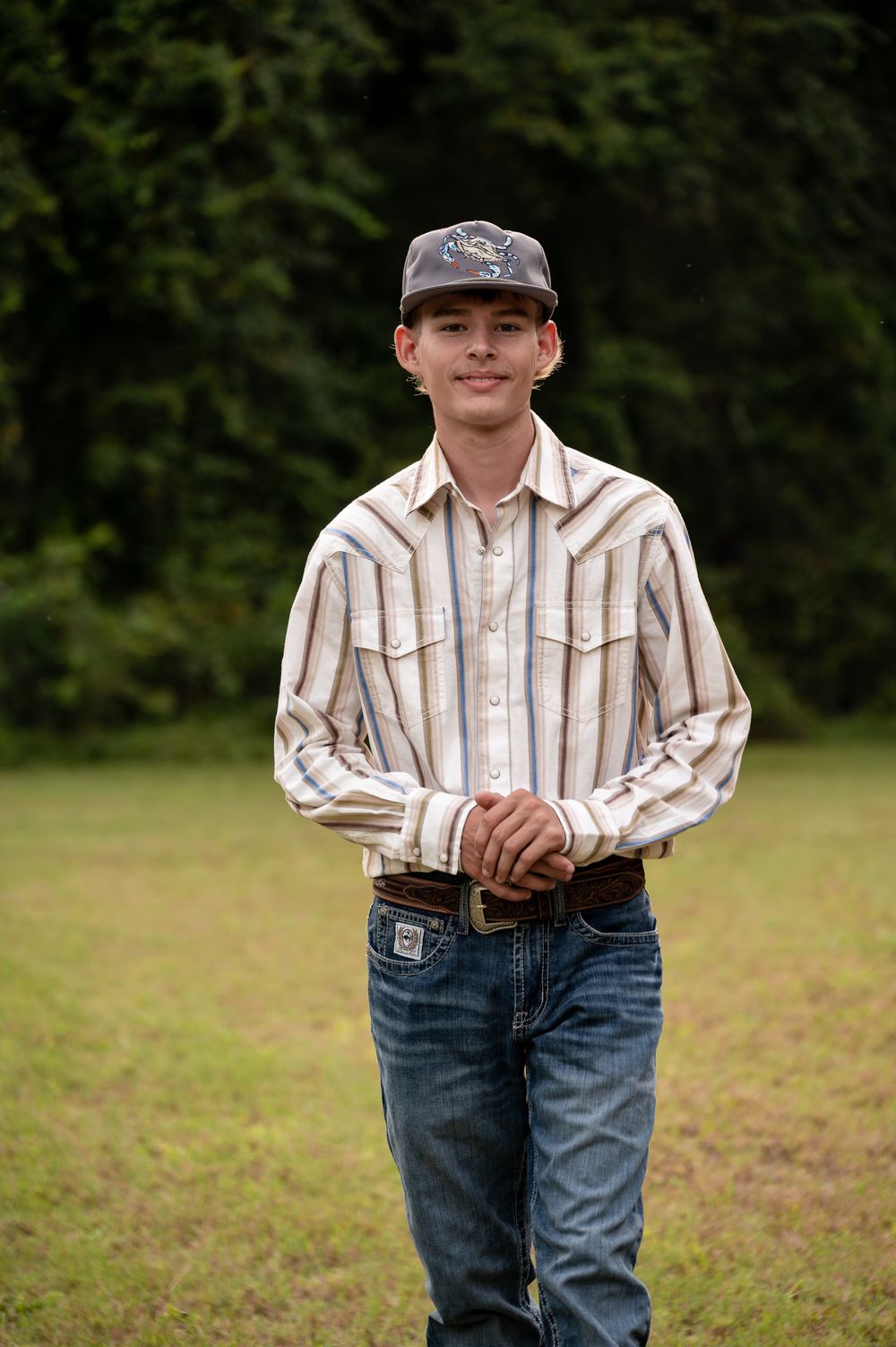 Young man in striped western shirt, jeans, and cap smiling outdoors on green grass with trees in background.