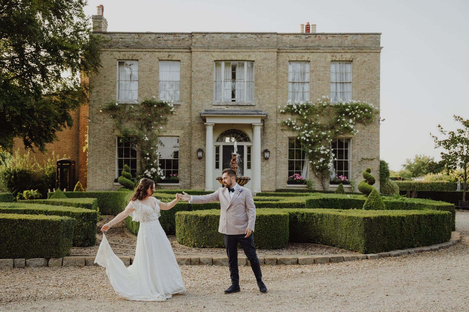 Bride and groom dance joyfully in front of elegant stone manor house surrounded by manicured hedges. Victoria, BC, Vancouver Island.