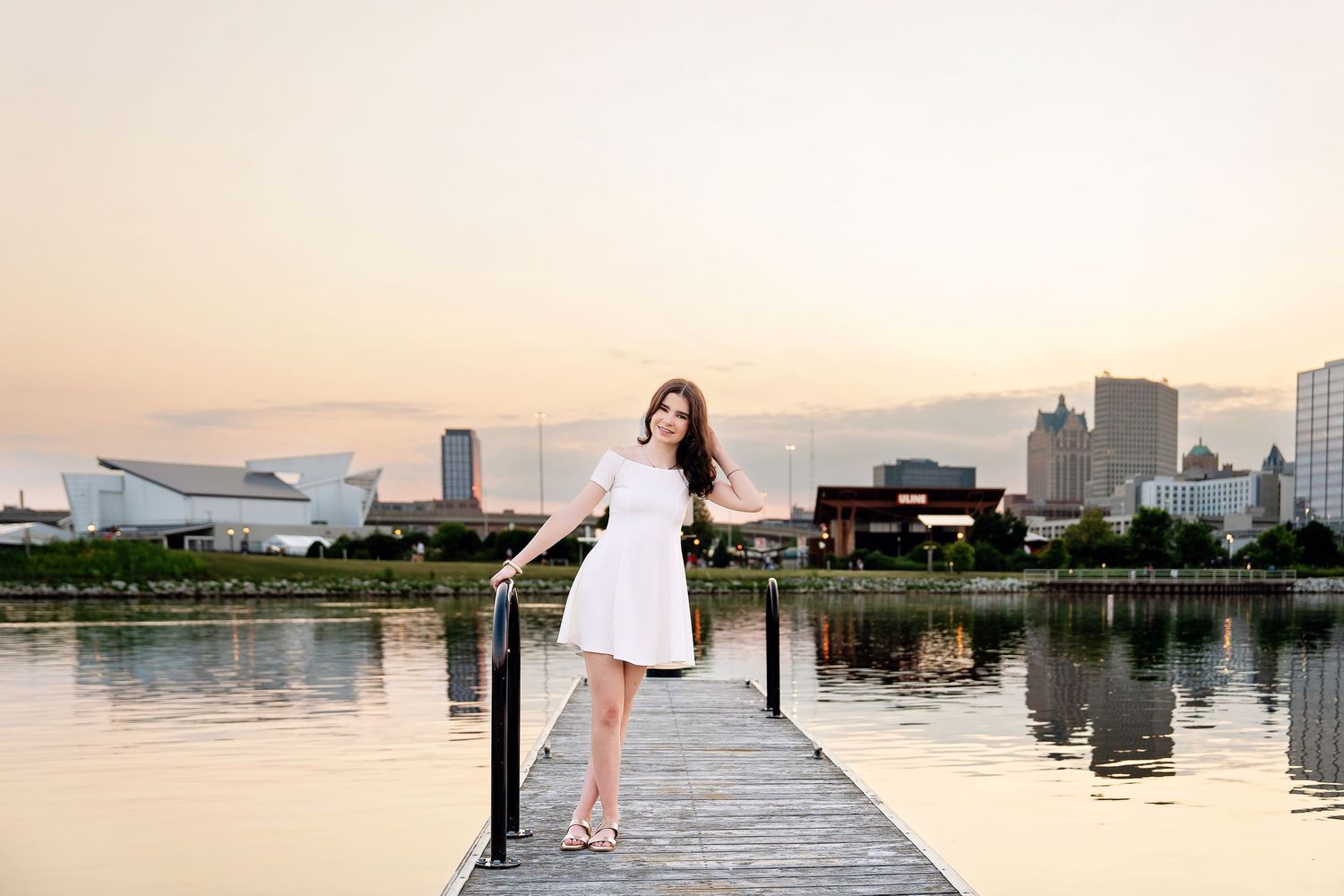Senior photo by Rachel Dickman of young woman with the city sunset behind her standing on a dock with water around her.