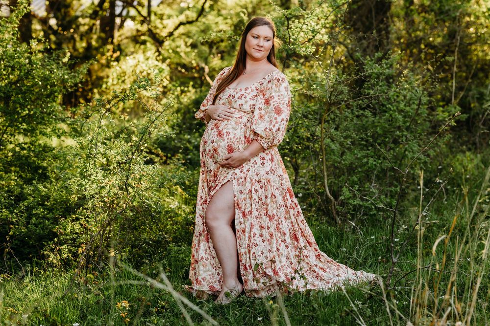 Pregnant woman in floral maxi dress posing outdoors in lush green nature setting for maternity photo shoot.