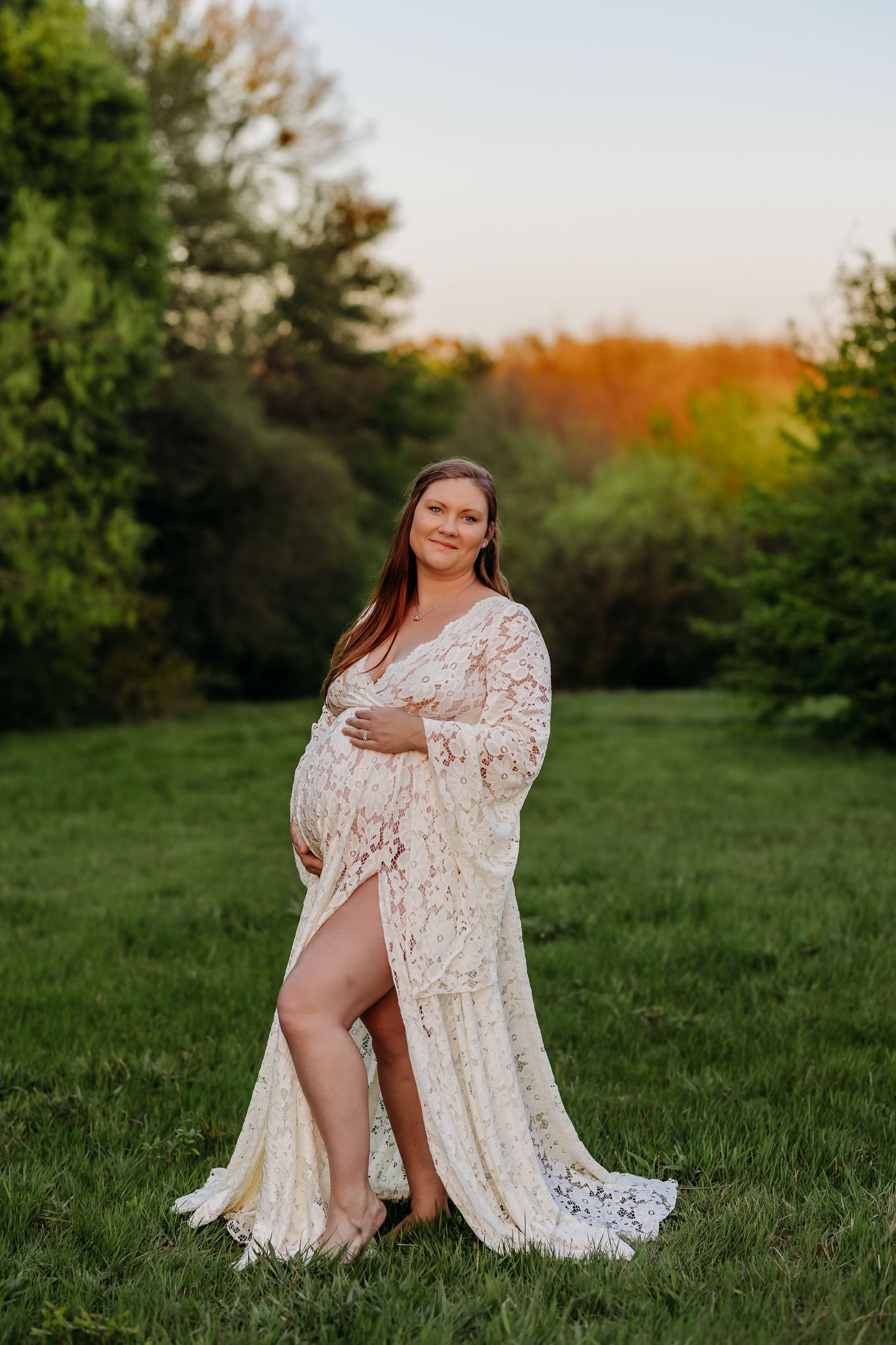 Pregnant woman in white lace maternity gown posing outdoors on green grass at golden hour sunset.