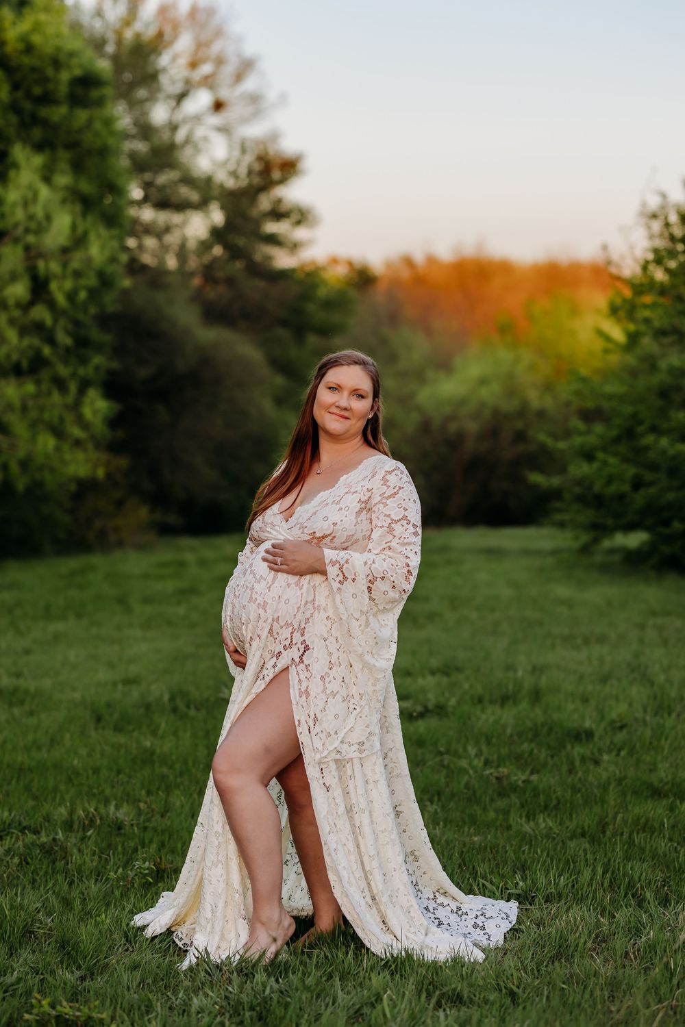 Pregnant woman in white lace maternity gown posing outdoors on green grass at golden hour sunset.