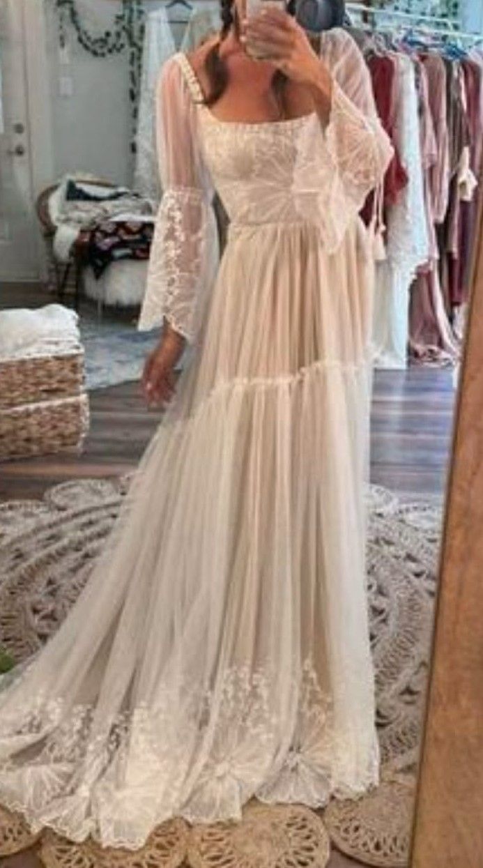 Woman modeling a boho white lace wedding gown with long sleeves and flowing tulle skirt in a bridal boutique.