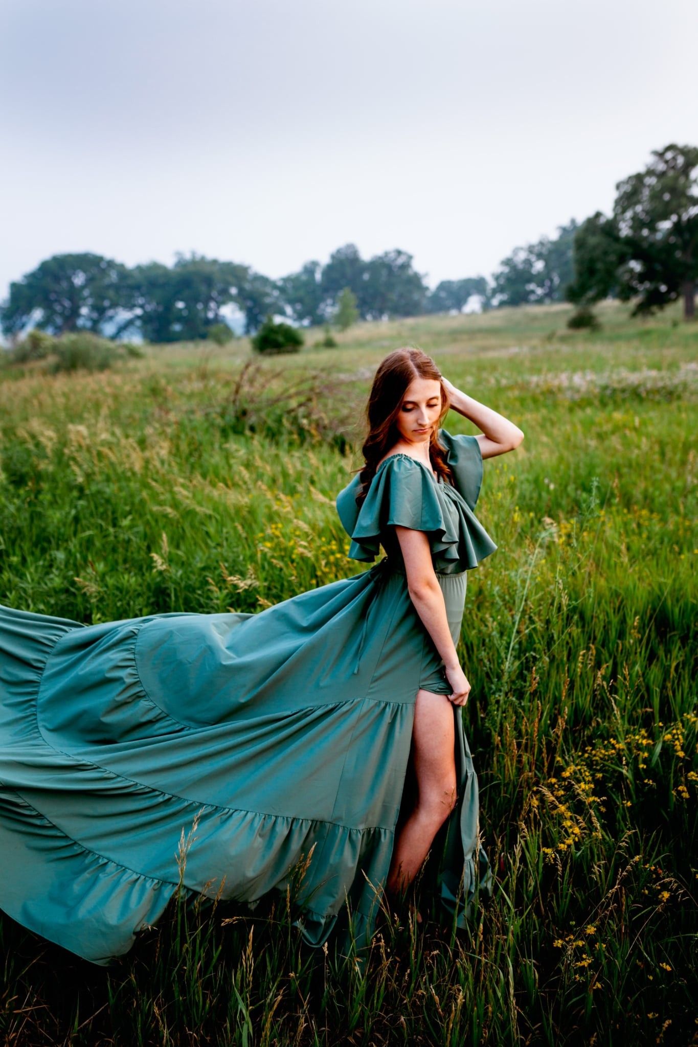 A flowing teal dress creates a dramatic silhouette against a rustic meadow landscape in summer.