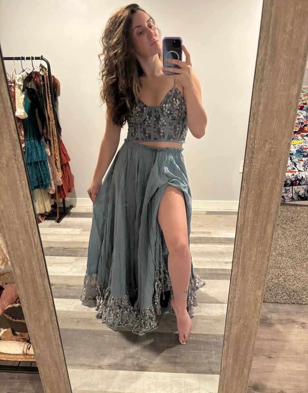 A figure poses in front of a mirror wearing a flowy gray dress with lace trim details in a fitting room.
