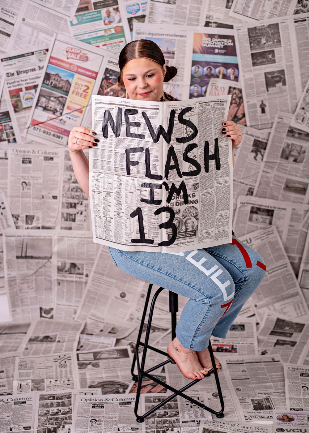 Girl holds newspaper reading News Flash I'm 13, seated on stool with newspaper backdrop, birthday photo shoot.