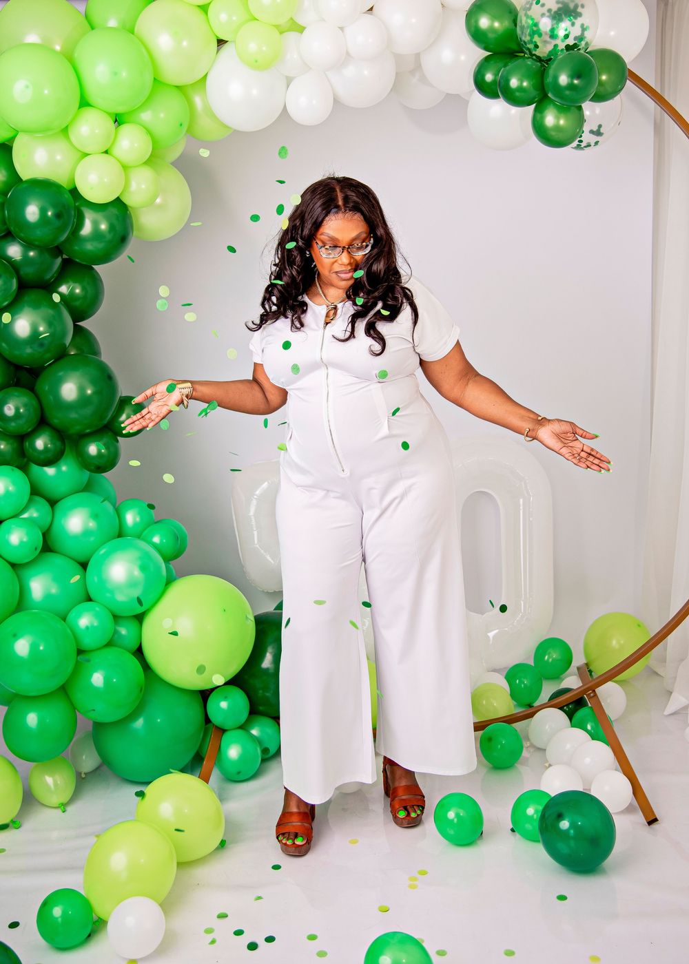 Woman in white jumpsuit posing with green and white balloon arch, tossing confetti in a celebratory photo shoot.