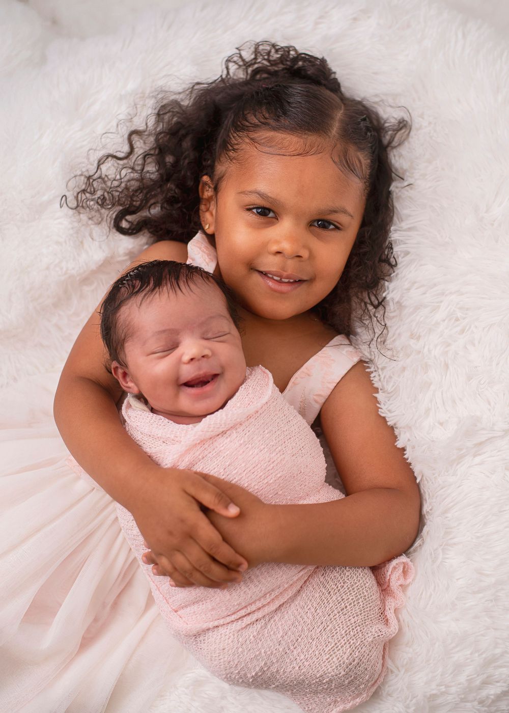A smiling young girl with curly hair holds a sleeping newborn baby wrapped in a pink swaddle on a white fluffy blanket.