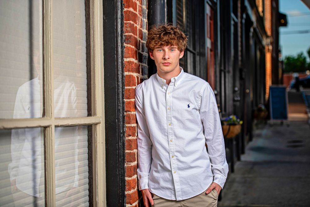 Young man in white button-up shirt leaning against brick wall on urban sidewalk for senior portrait.