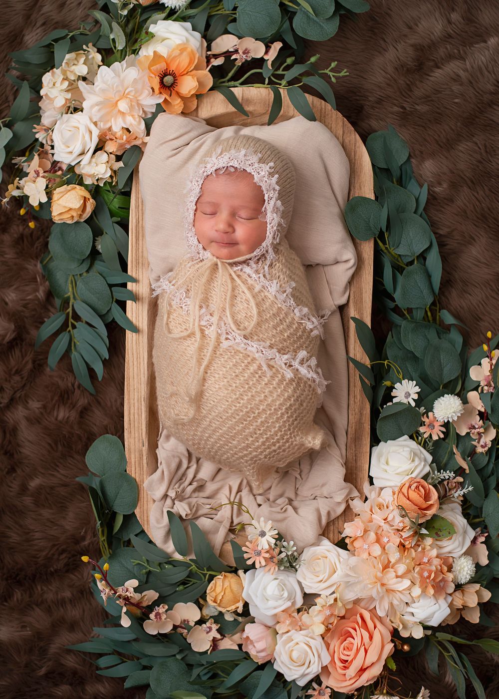 Sleeping newborn swaddled in cream knit wrap and lace bonnet, nestled in wooden bowl surrounded by peach and white florals.