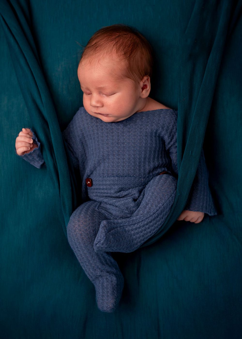 Sleeping newborn baby in blue knit outfit posed on teal fabric backdrop in professional newborn photography.