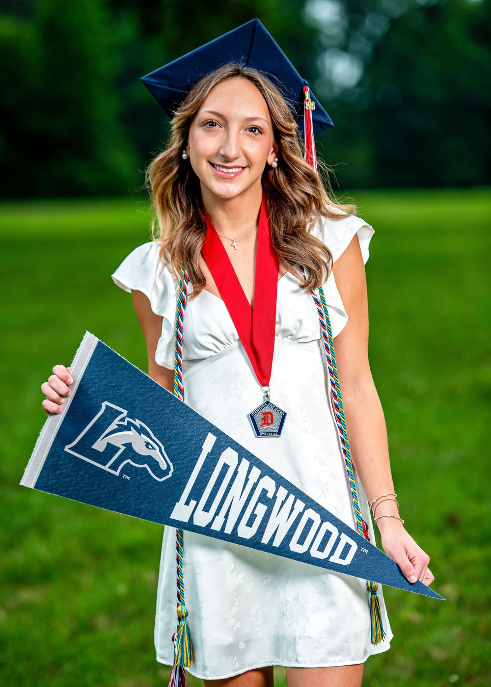 Smiling graduate in cap and gown holds Longwood University pennant and wears medal outdoors.