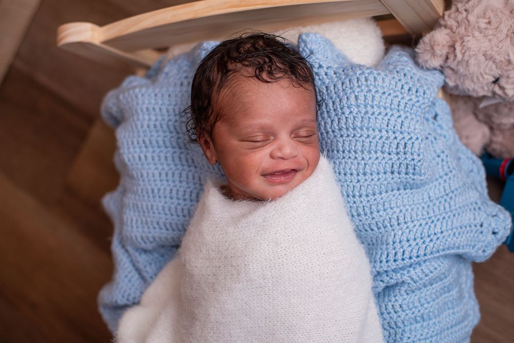 Smiling newborn baby swaddled in white wrap on blue crochet blanket in wooden bassinet.