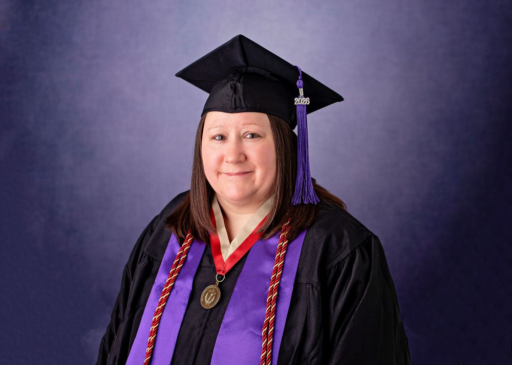 Woman in black graduation gown and cap with purple hood and gold medal smiles for formal graduation portrait.