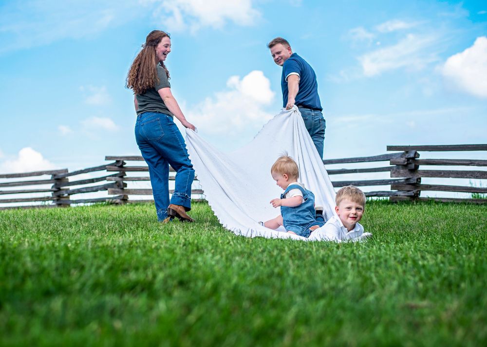Family spreading white blanket on green grass field with wooden fence and blue sky backdrop.