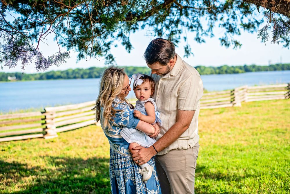 A young family with a baby girl poses outdoors near a lake under a tree on a sunny day.