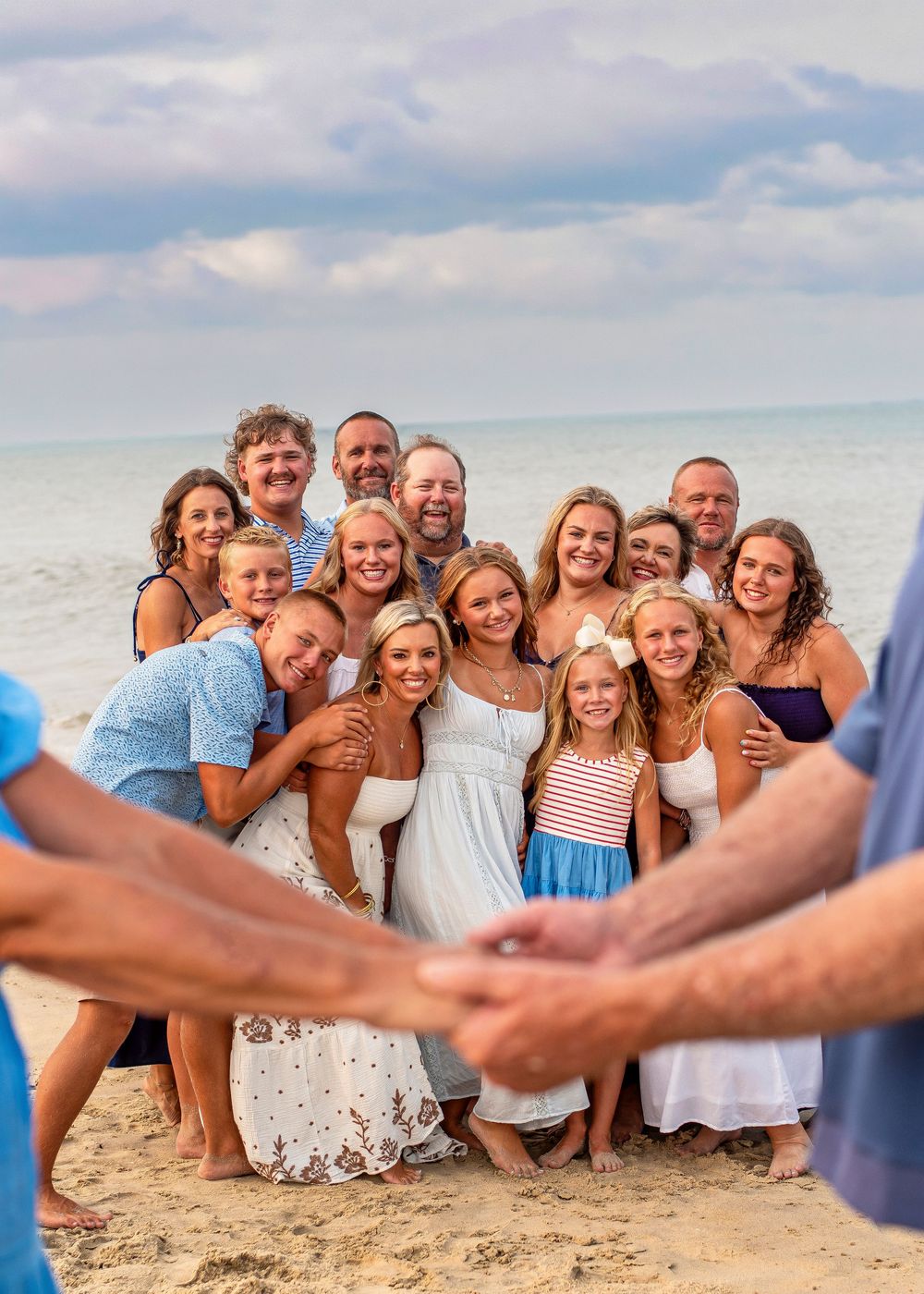 Large family group photo on a sandy beach with ocean backdrop, smiling adults and children posing together on a cloudy day.