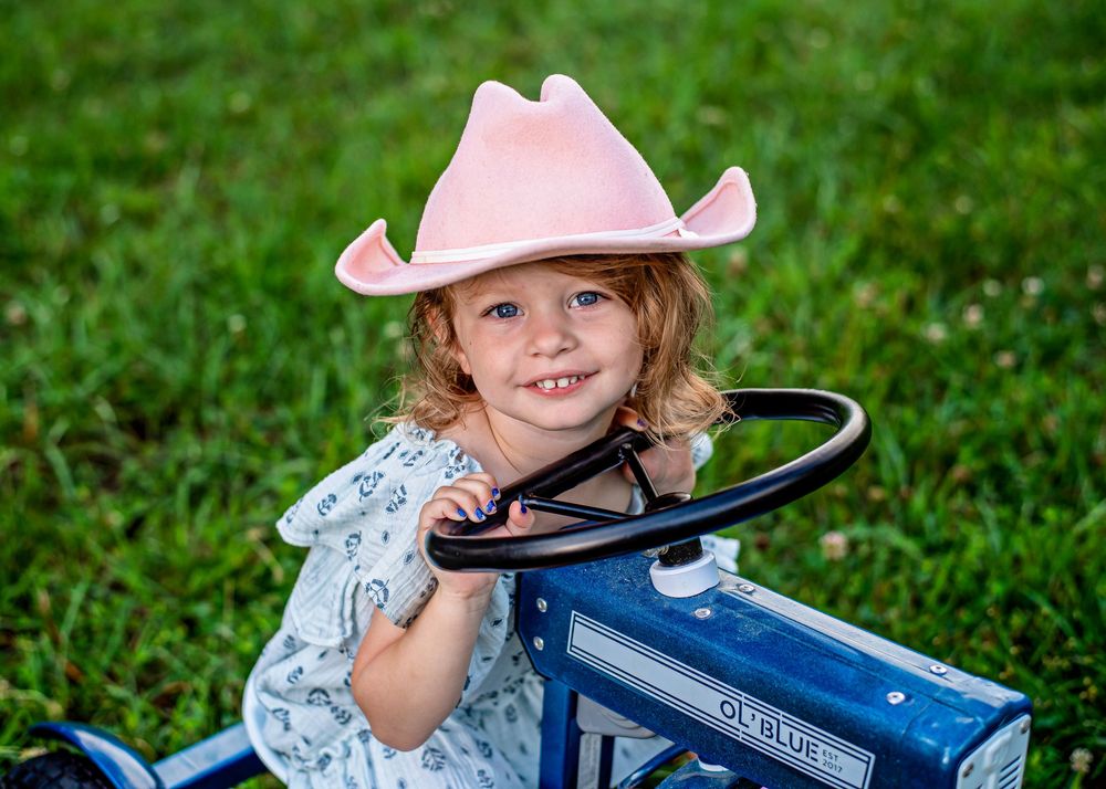 Smiling toddler girl in pink cowboy hat sitting on blue tractor on green grass.