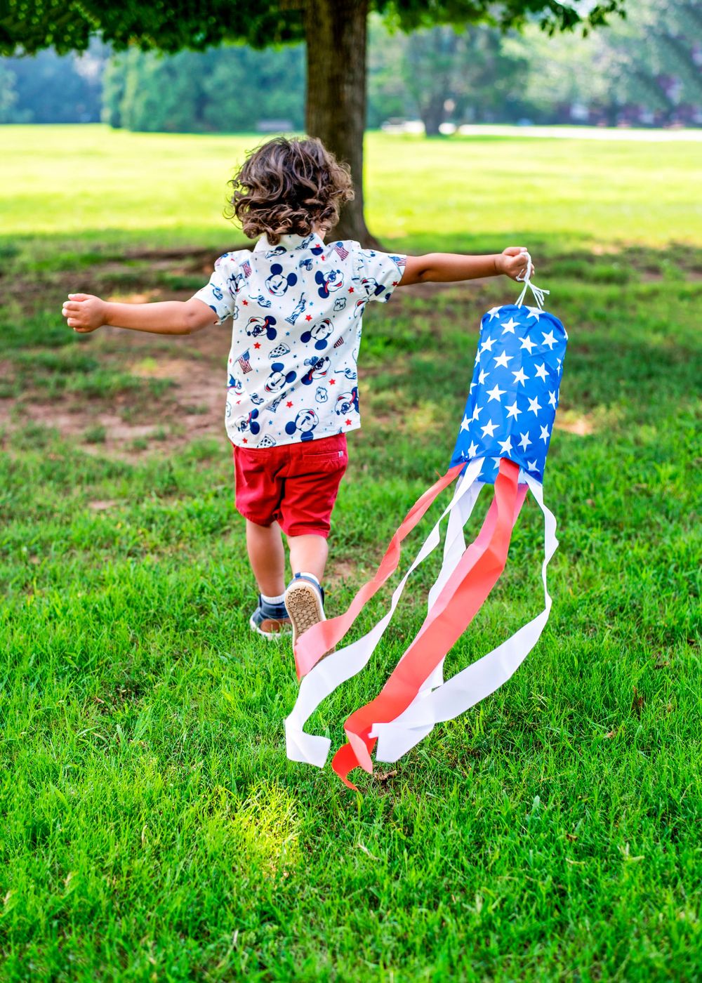 Child running on grass holding an American flag windsock, wearing a Mickey Mouse shirt and red shorts.