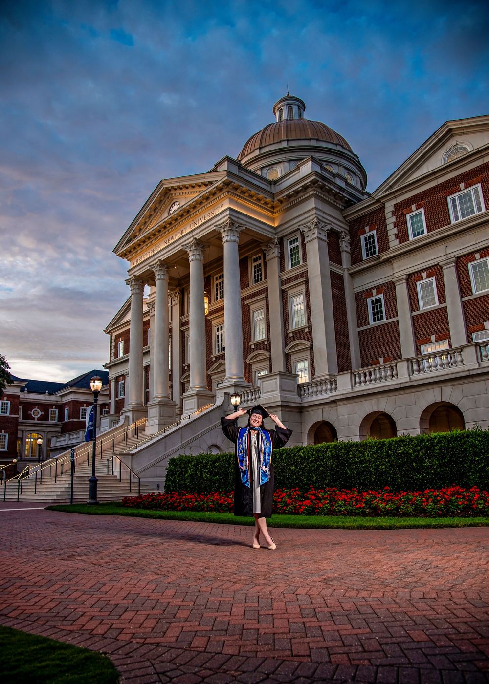 Graduate in cap and gown poses in front of a grand domed brick university building at dusk.