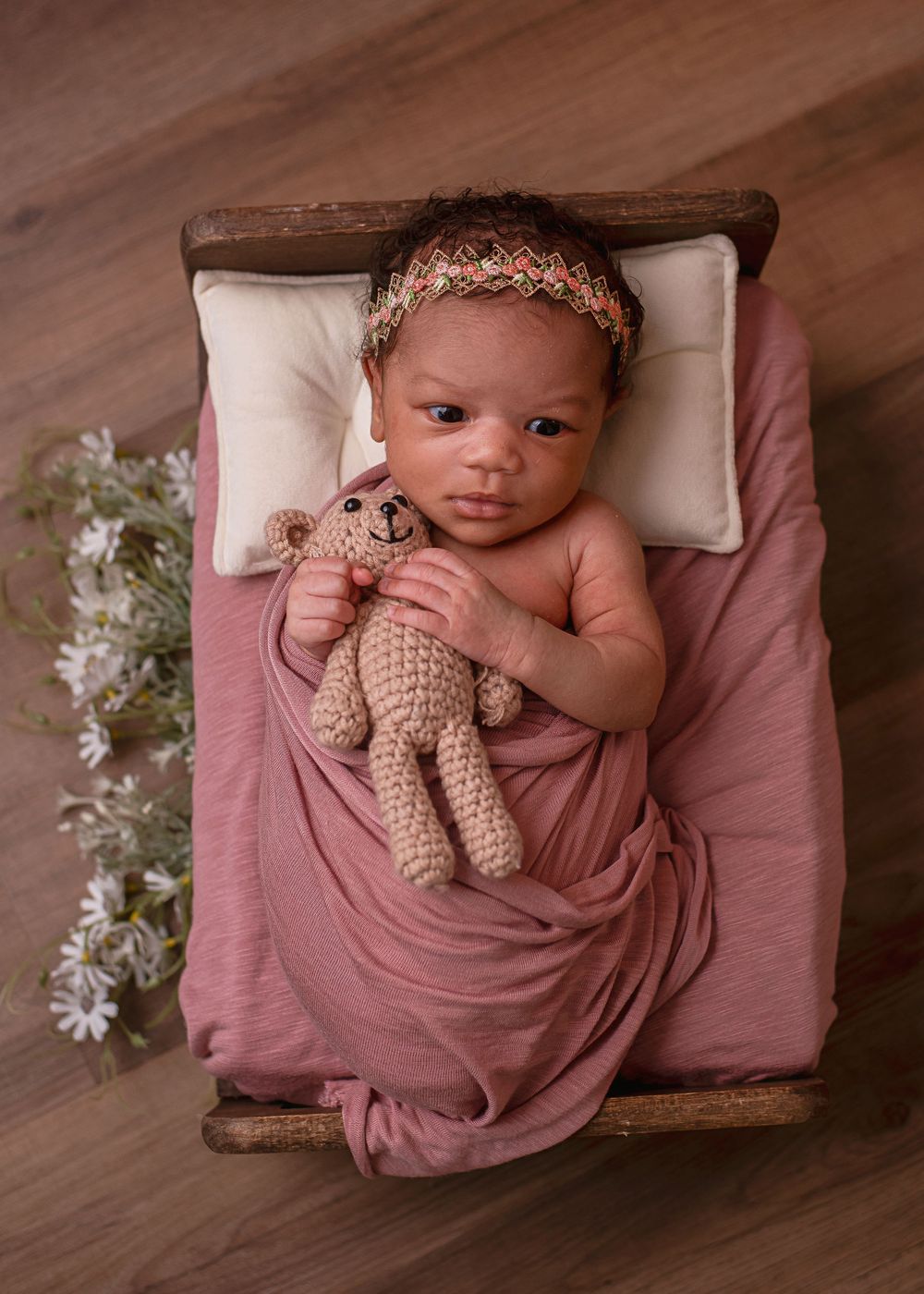 Newborn baby wrapped in pink swaddle holding crochet teddy bear on miniature wooden bed with floral headband.