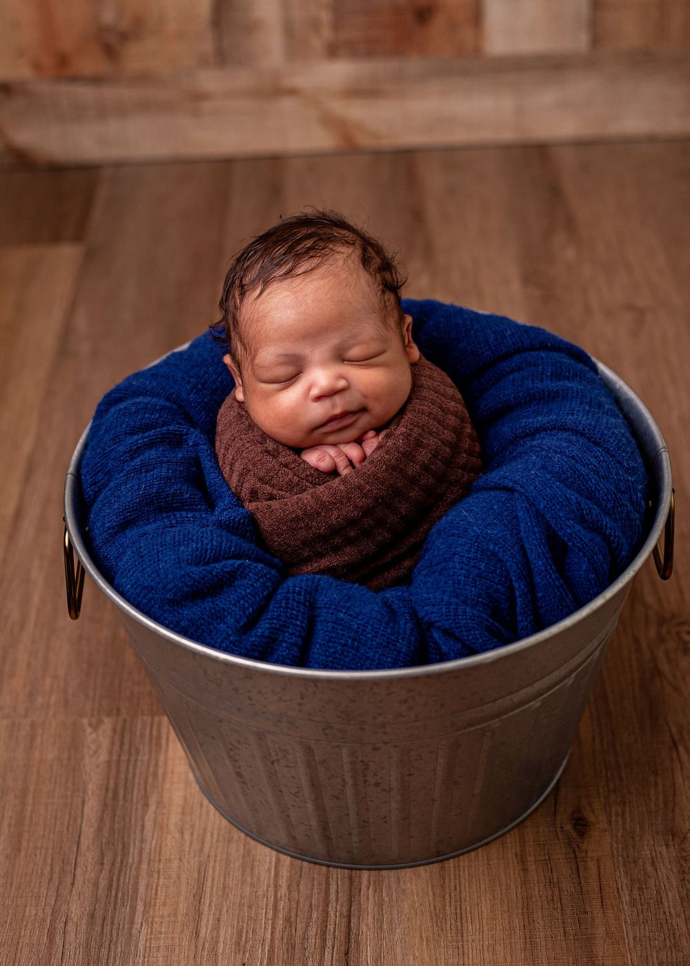 Sleeping newborn baby wrapped in brown knit swaddle nestled in blue blanket inside silver metal bucket on wood floor.