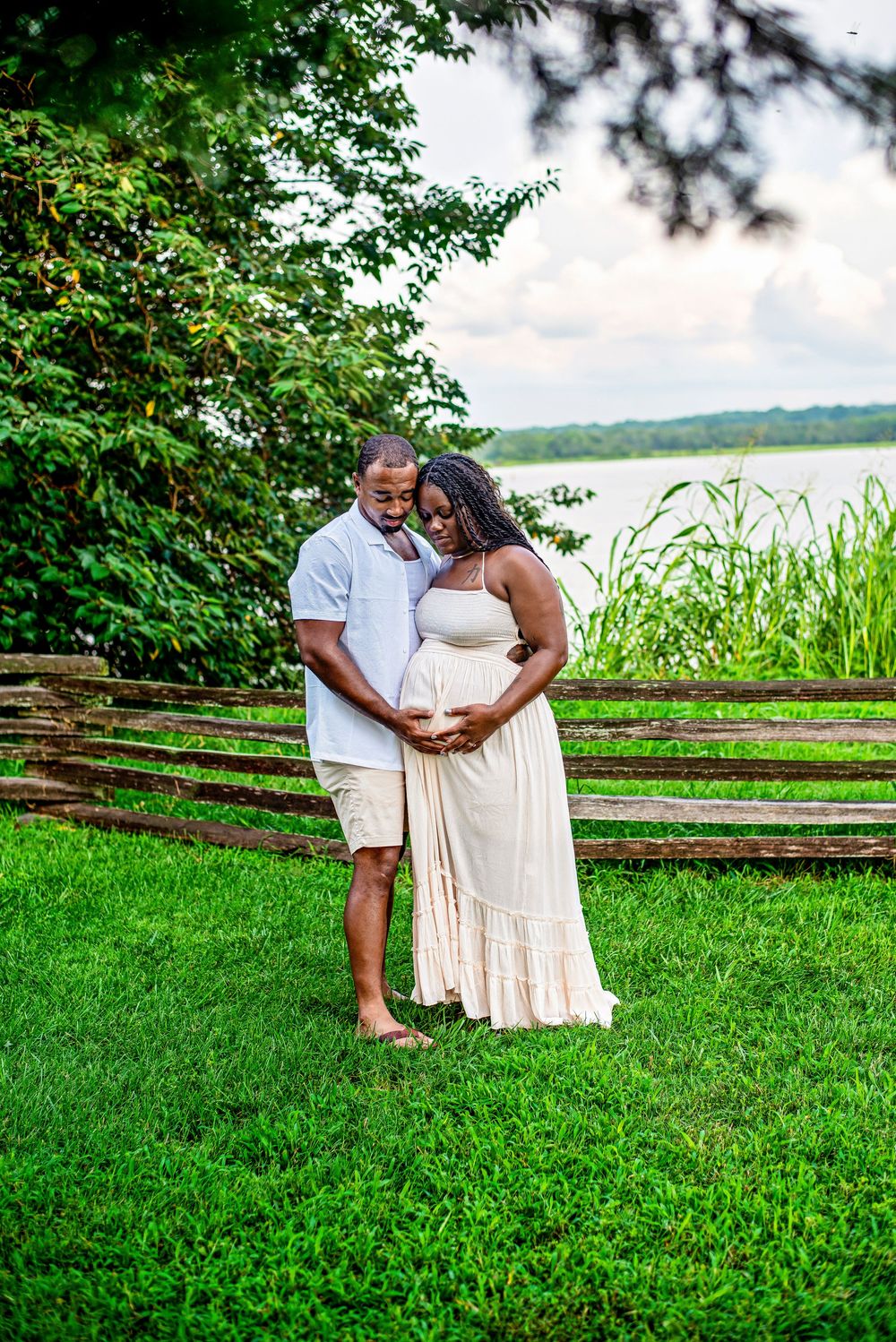Couple sharing a tender maternity moment outdoors by a lake, woman in white dress cradling baby bump.