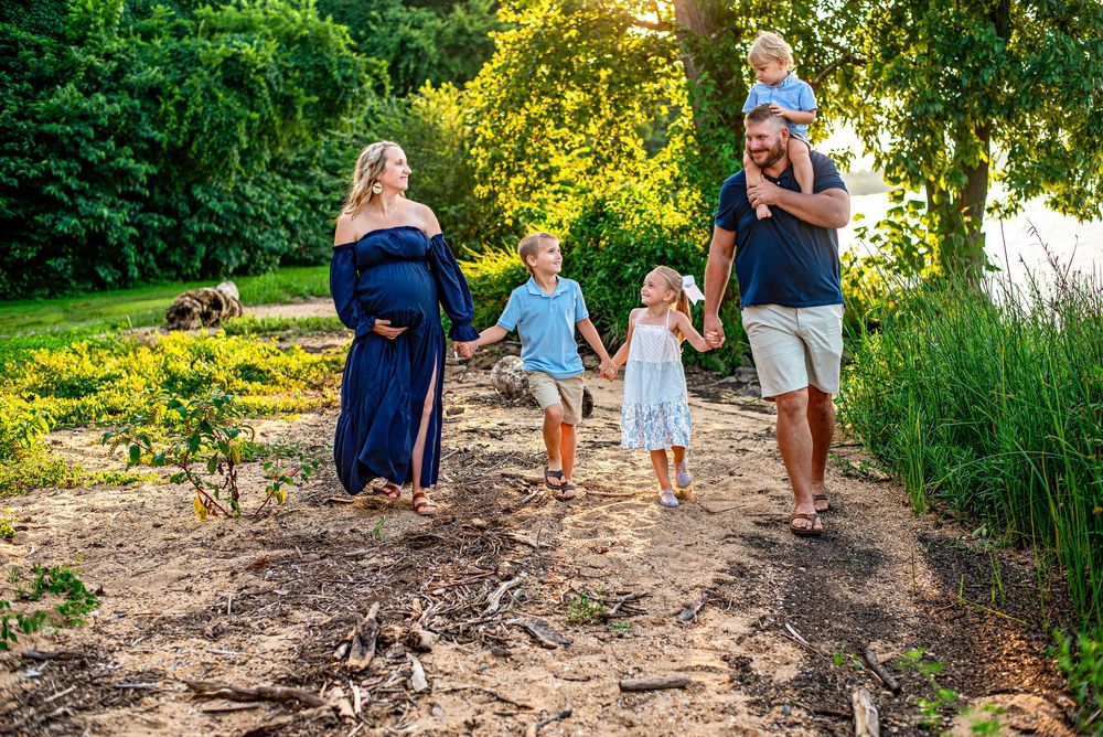 Pregnant woman in navy dress walks with family including three children and husband in sunny green garden path.