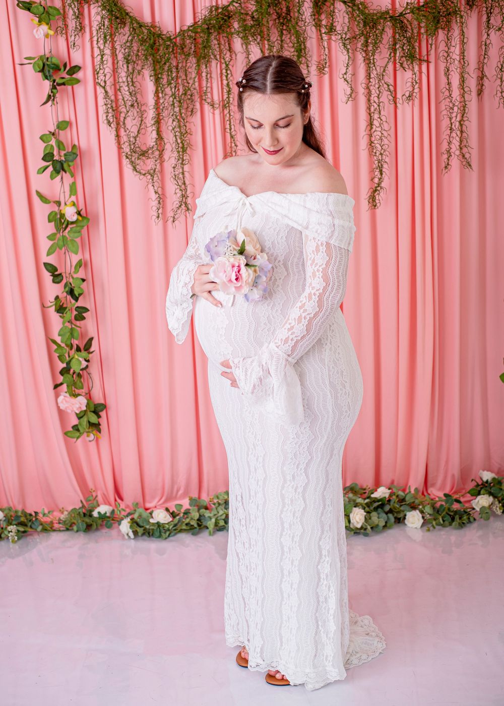 Pregnant woman in white off-shoulder lace gown holding flowers, posing before pink backdrop with greenery.