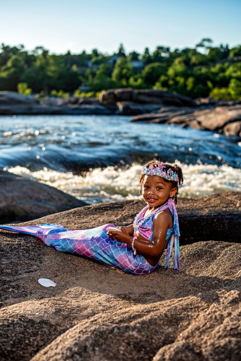 Young girl in colorful mermaid costume with tiara sits on rocks near rushing river, smiling at camera.