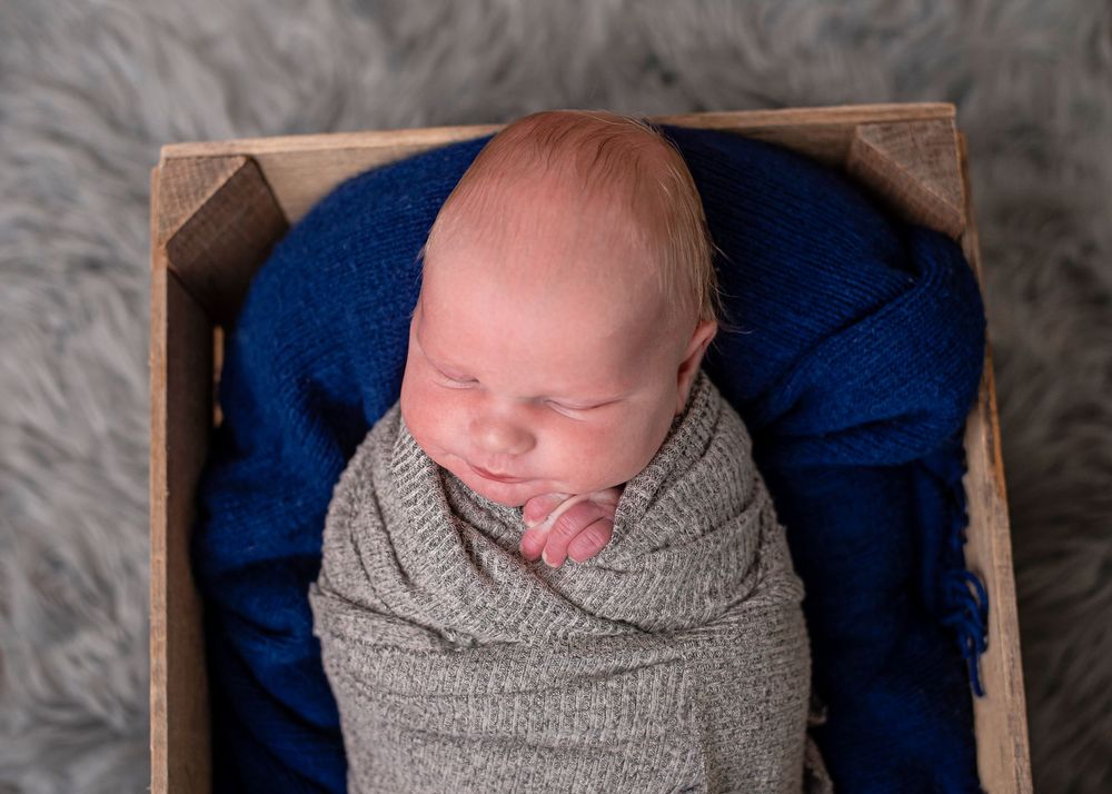 Sleeping newborn baby swaddled in gray wrap inside a rustic wooden crate with blue fabric backdrop.