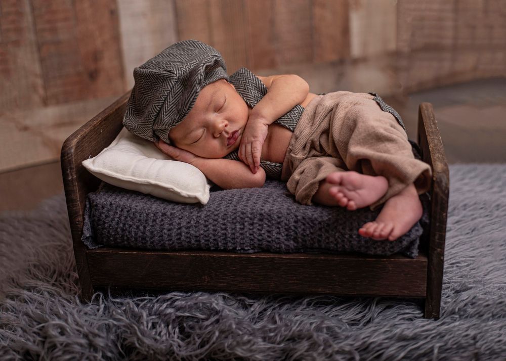 Newborn baby sleeping peacefully on a tiny wooden bed with gray bedding, wearing a knit hat and suspenders outfit.