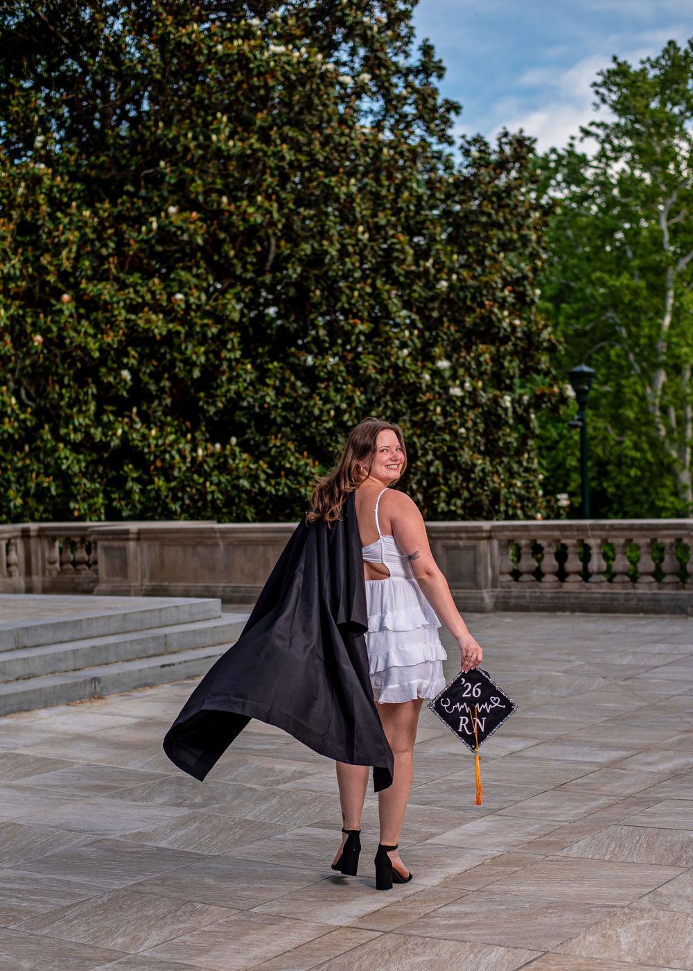 Nursing graduate in white dress holds black cap reading RN 26, posing outdoors with graduation gown flowing.