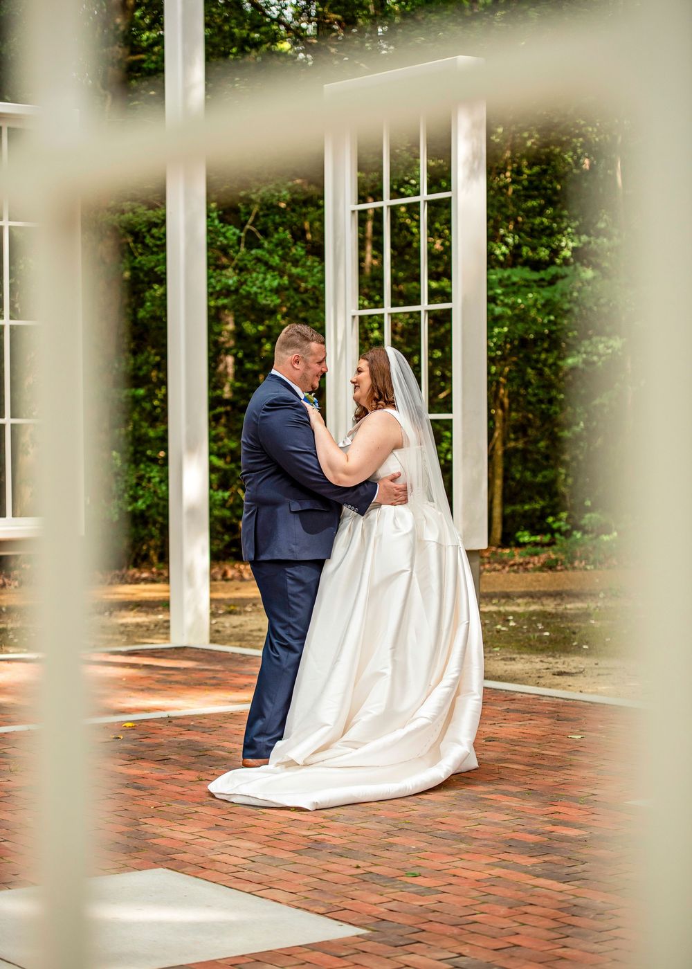 Bride and groom share a romantic embrace on a brick patio surrounded by white columns and lush greenery.
