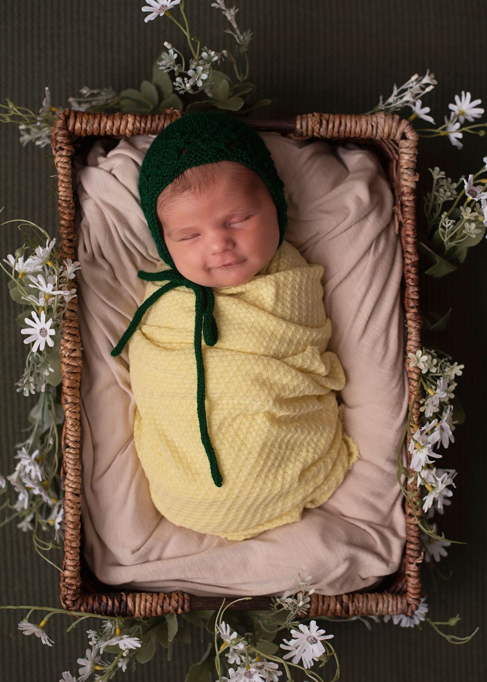 Sleeping newborn swaddled in yellow wrap wearing green knit bonnet, nestled in wicker basket with white flowers.