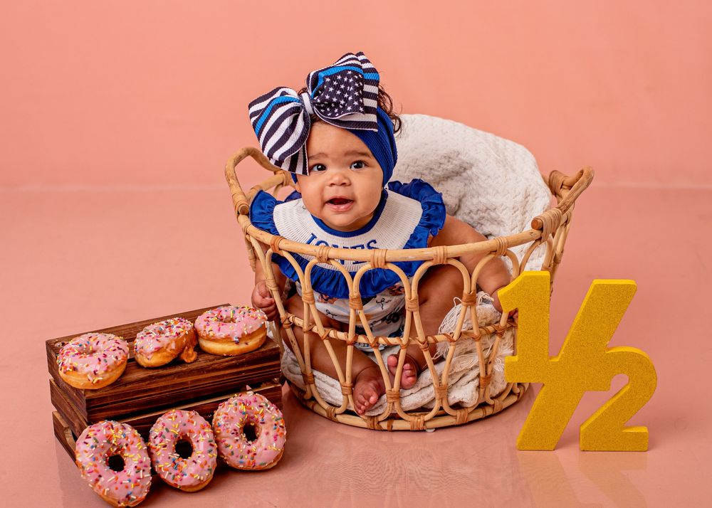 Baby girl in blue outfit sits in wicker basket with donuts and half birthday sign for 6-month milestone photo.