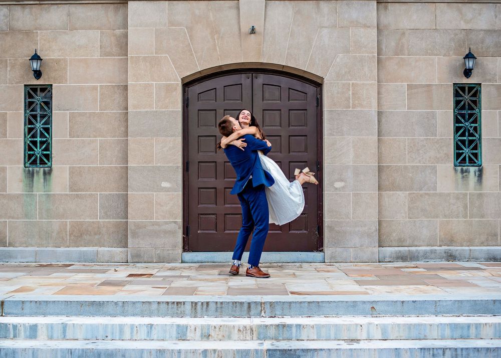Groom in navy suit lifting bride in white dress outside elegant stone building with arched wooden doors.