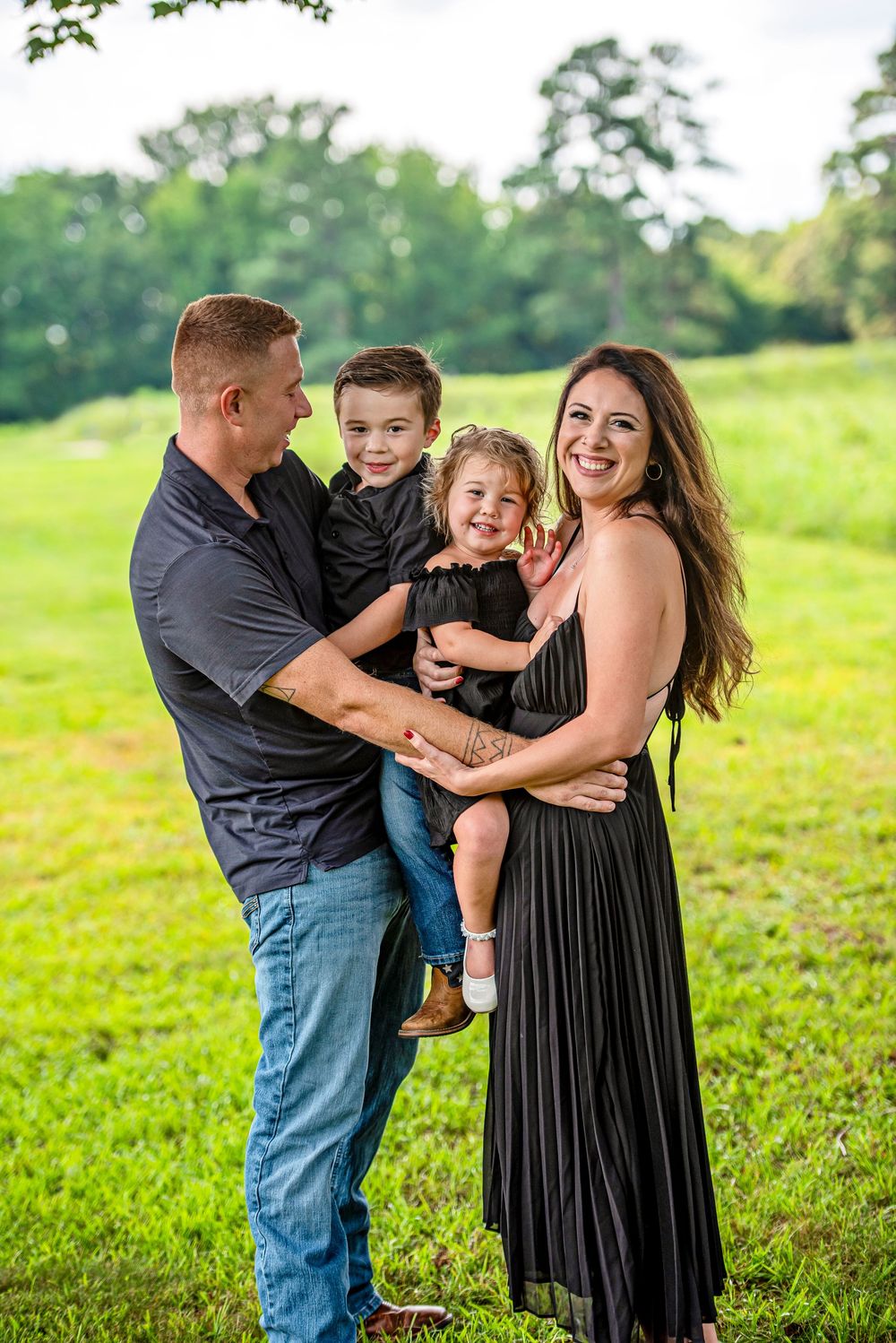 Happy family of four posing outdoors on green grass, parents holding two young children, all wearing coordinating dark outfits.