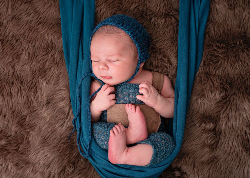 Newborn baby sleeping in a teal fabric hammock, wearing a blue knit bonnet, on a brown fluffy rug.