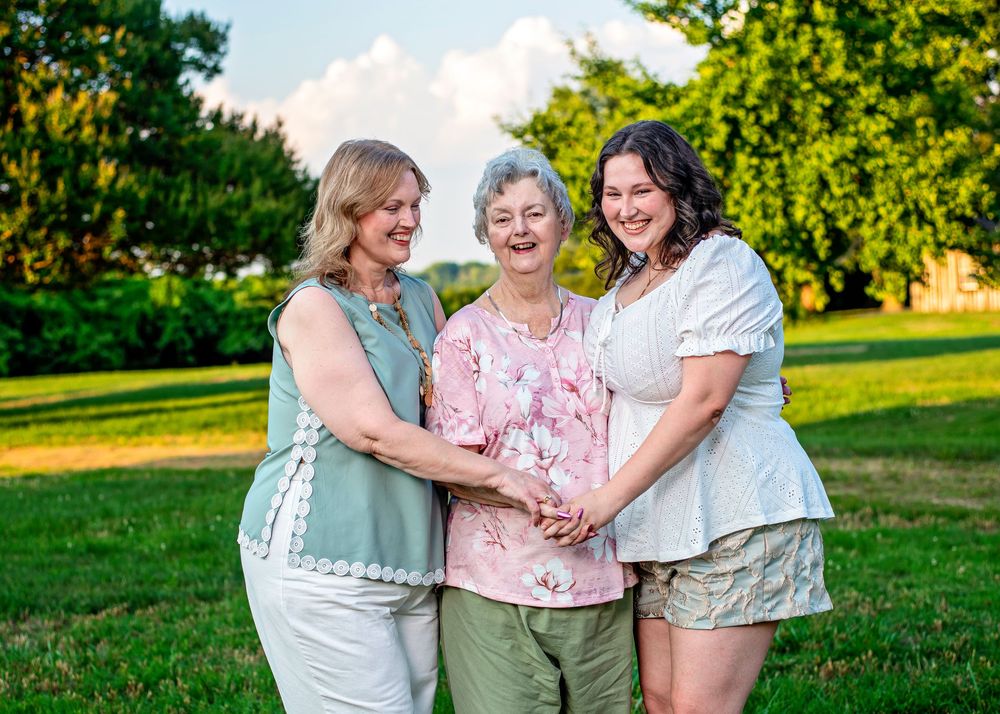 Three women smiling and posing together outdoors on green grass with trees in the background on a sunny day.
