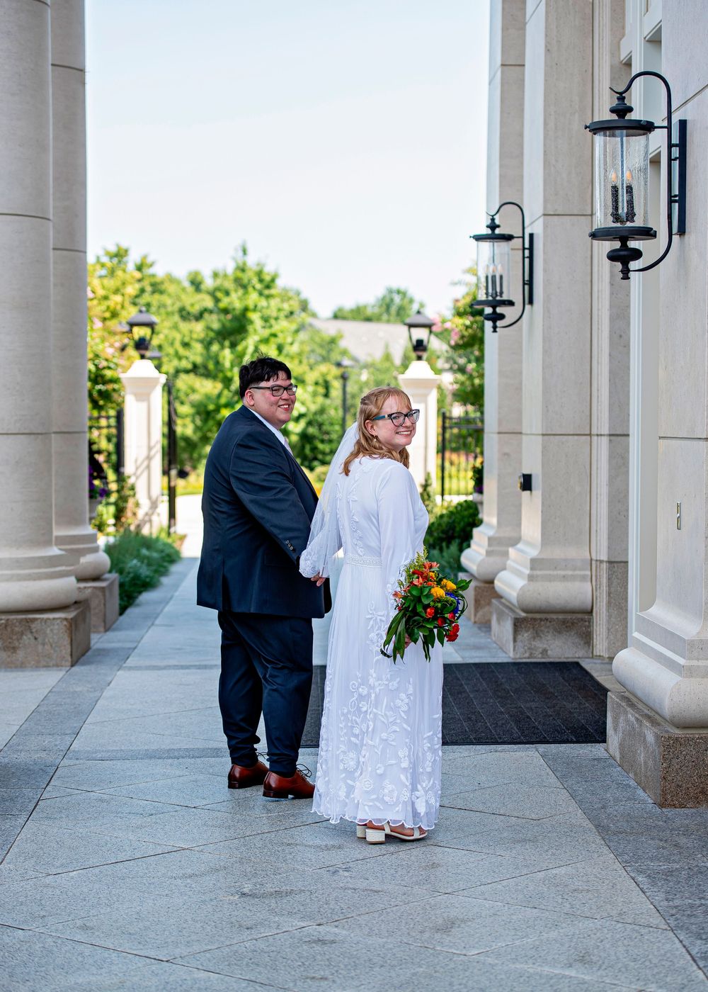 Couple in wedding attire holding a colorful bouquet pose on elegant stone columns pathway outdoors.