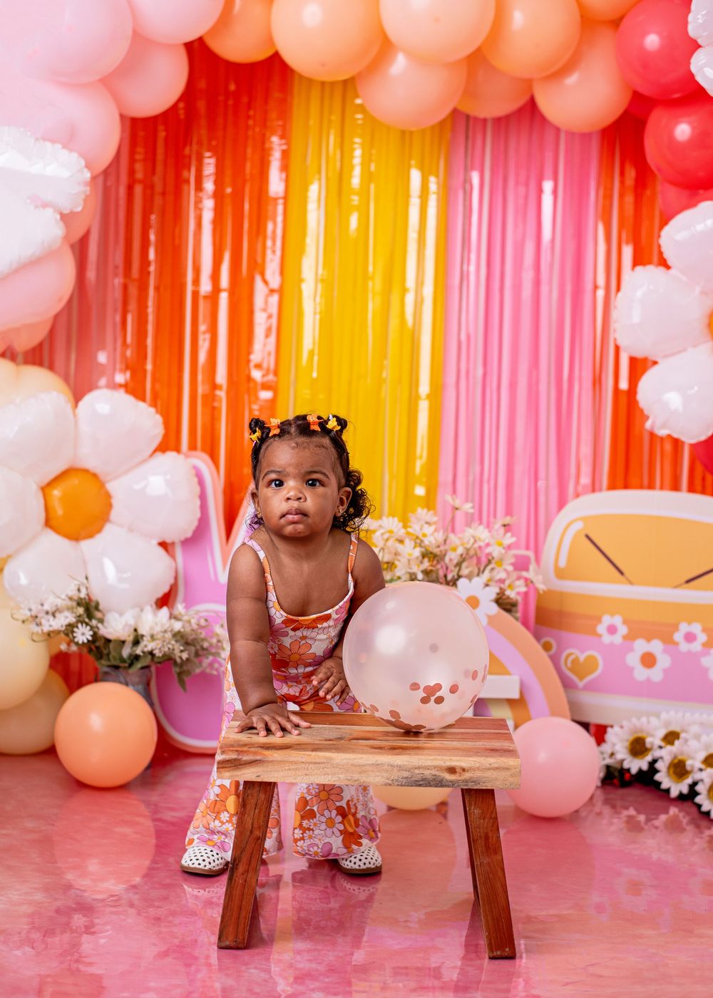 Toddler girl in floral dress sits at wooden stool surrounded by colorful balloons and daisy decorations at a birthday party.
