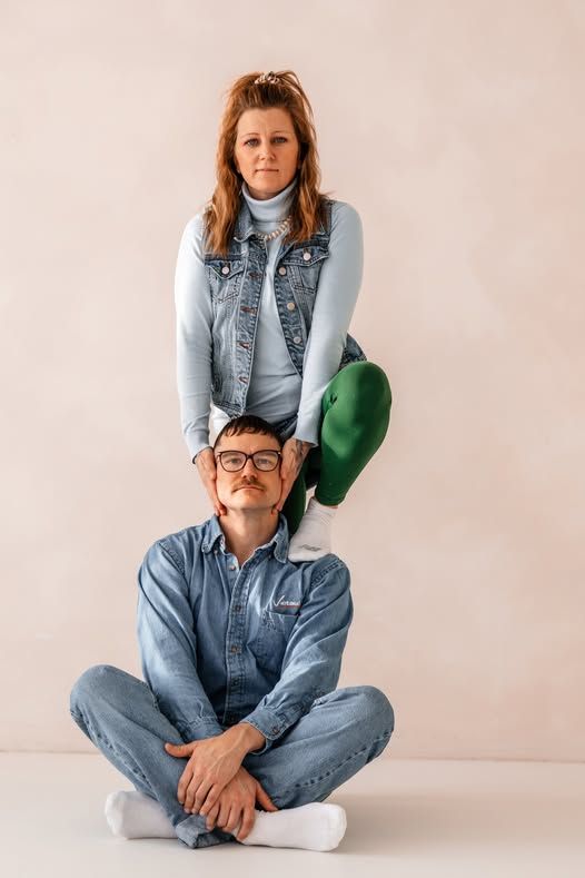 A man in a denim jumpsuit sits cross-legged on the floor while a woman in a denim vest stands behind him holding a green bag.