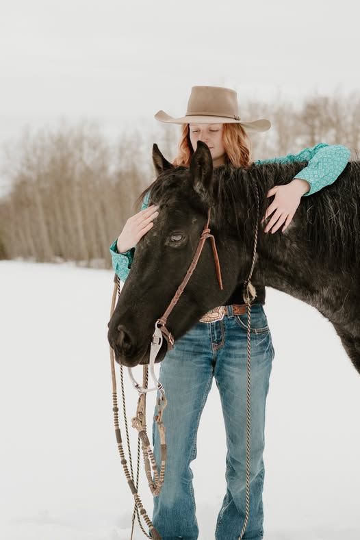 A cowgirl in a tan hat and turquoise shirt tenderly embraces a black horse in a snowy winter landscape.