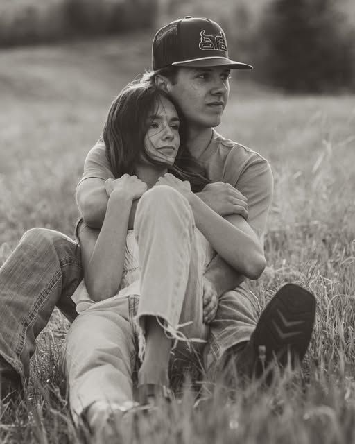 Black and white photo of a young couple sitting in a grassy field, the man embracing the woman from behind.