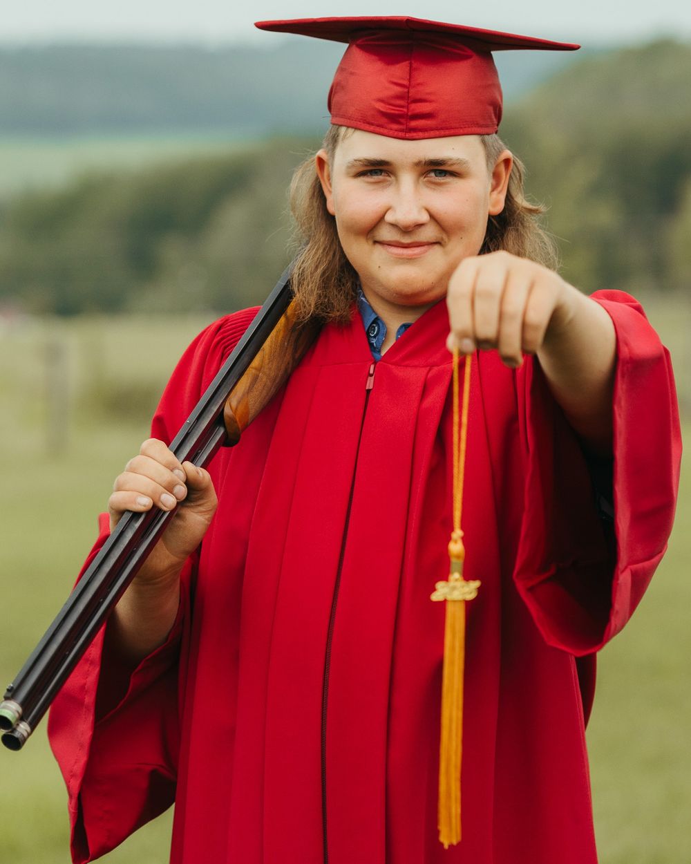 Graduate in red cap and gown holding a shotgun and gold tassel outdoors.