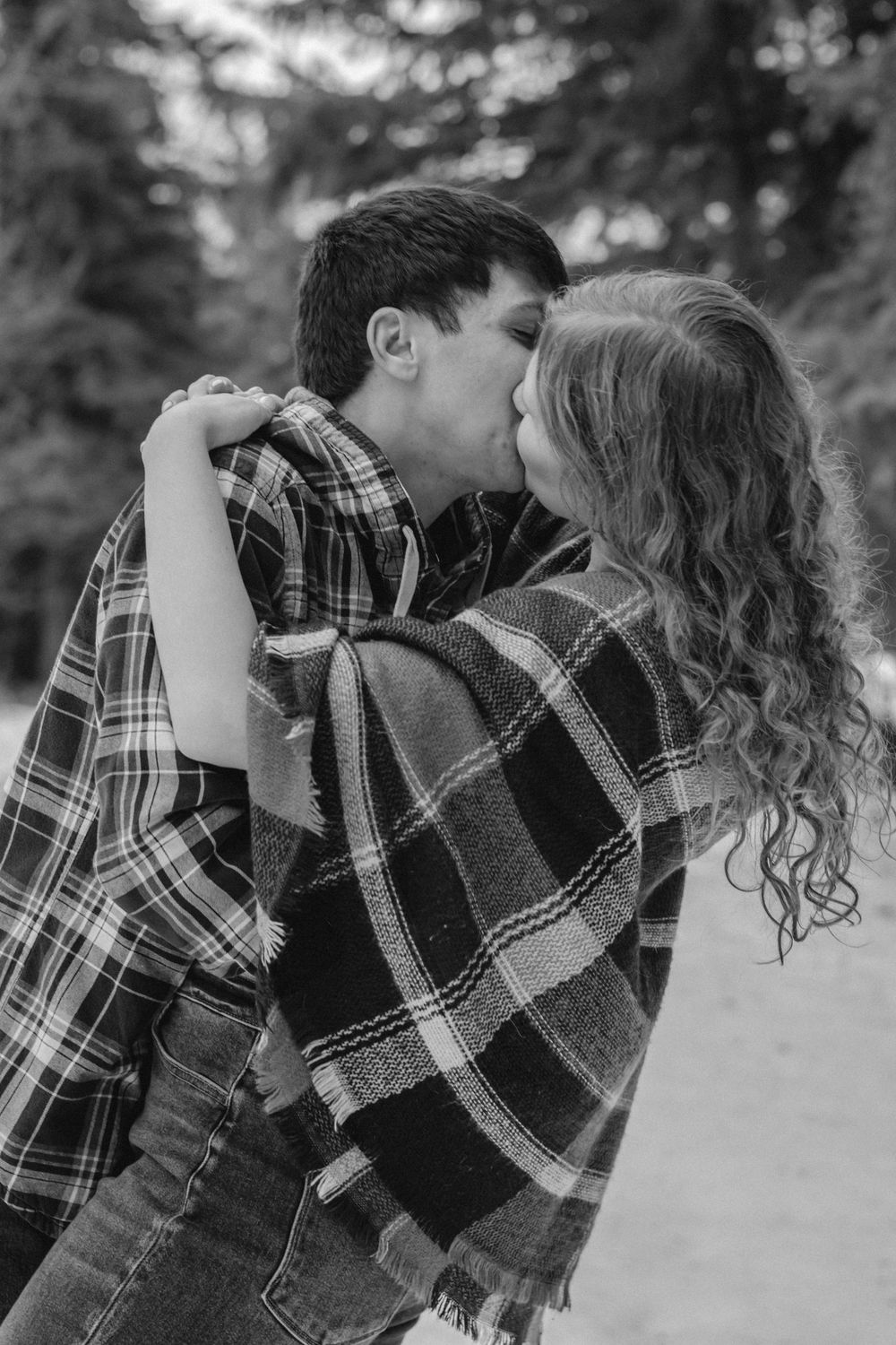 Black and white photo of a couple sharing a romantic kiss while wrapped in a plaid blanket outdoors in winter.
