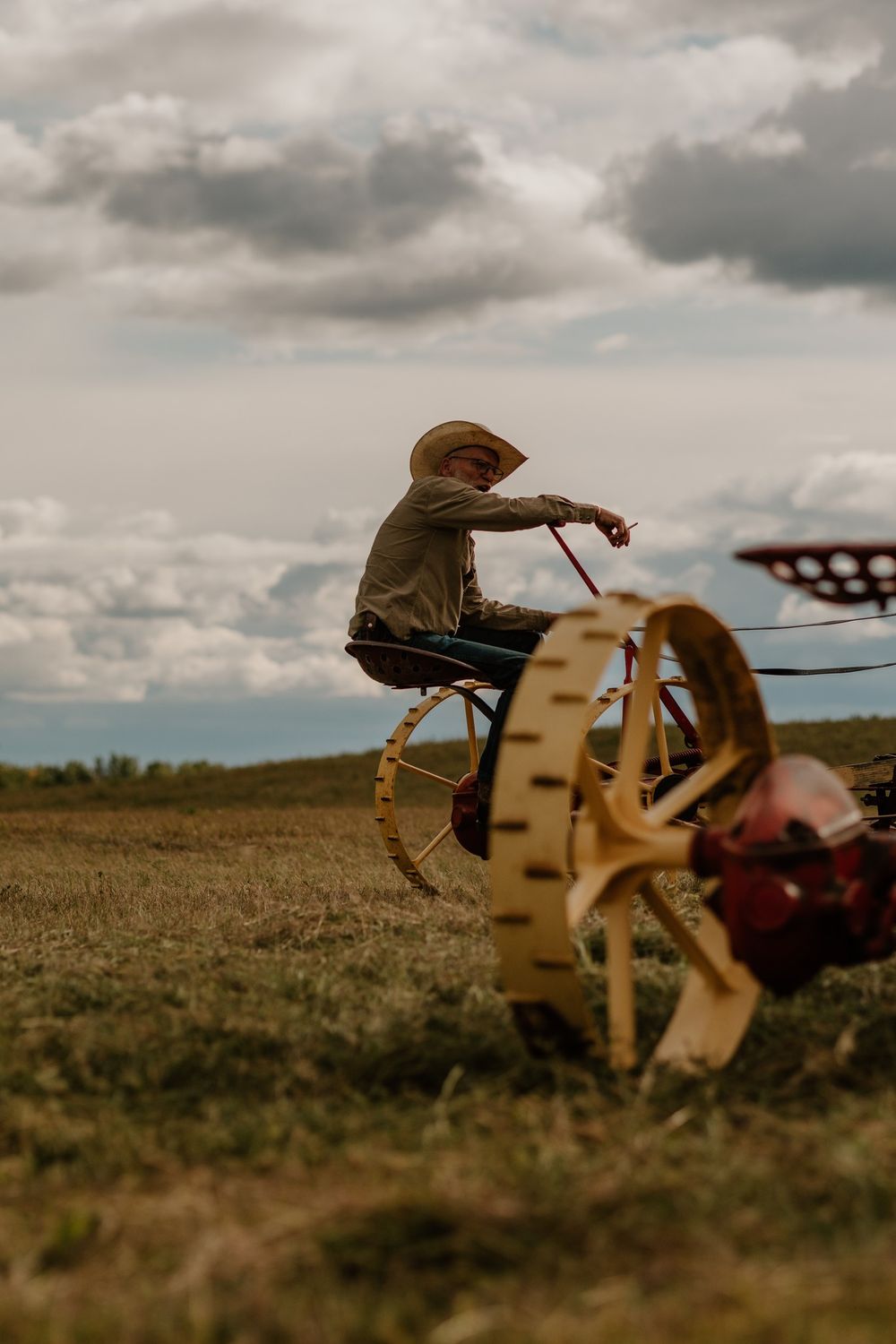 Farmer in a straw hat operating vintage horse-drawn plow equipment across an open field under dramatic cloudy skies.