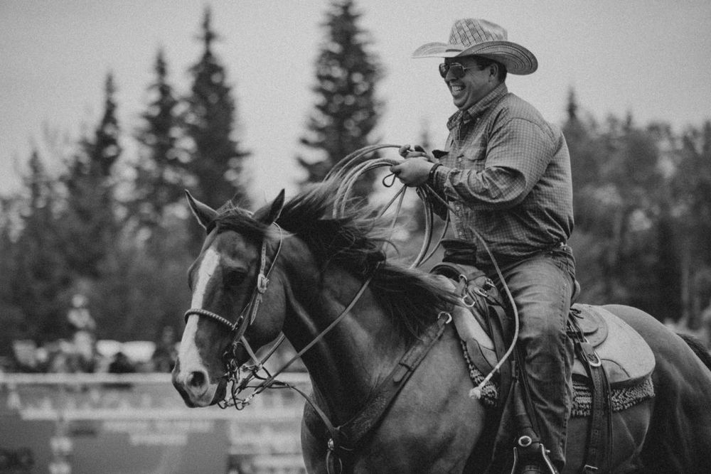 Cowboy in hat rides dark horse at outdoor rodeo event, black and white photography.