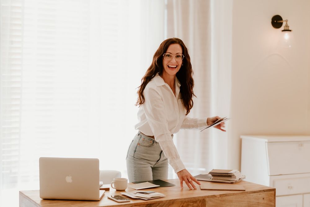 A smiling brunette woman in a white shirt leans over a wooden desk with a laptop and notebooks in a bright office.